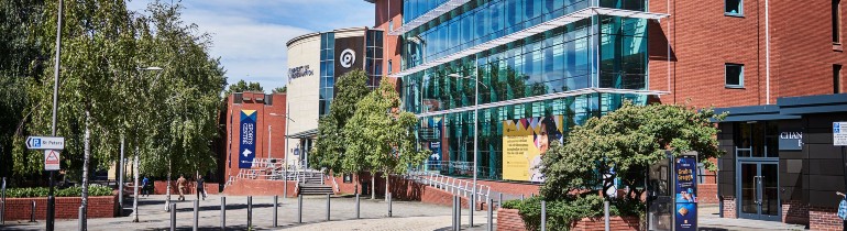 Exterior of the Ambika Paul building on a sunny day, a red-brick building with a large wall of windows currently decorated with a large graphic of a student.
