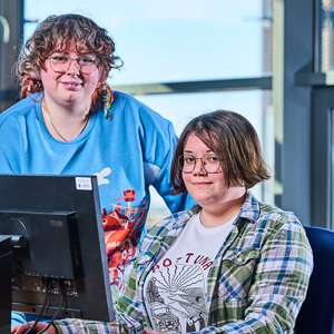 Two female students on campus at the student PCs. One sits at desk, the other, smiling is leaning over.