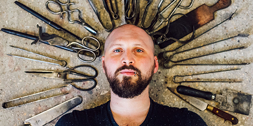 Photograph of Elliot Walker whose head is surrounded by a circle of bladed instruments: scissors, tongs and knives