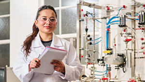 student wearing white lab coat and safety glasses, standing holding a clipboard in a chemical engineering lab at City campus.