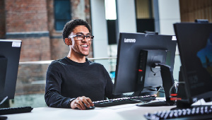 Student sat at desk working on computer and smiling. They are wearing glasses and have braces