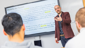 Teacher stood at front of class explaining to students what is on the board