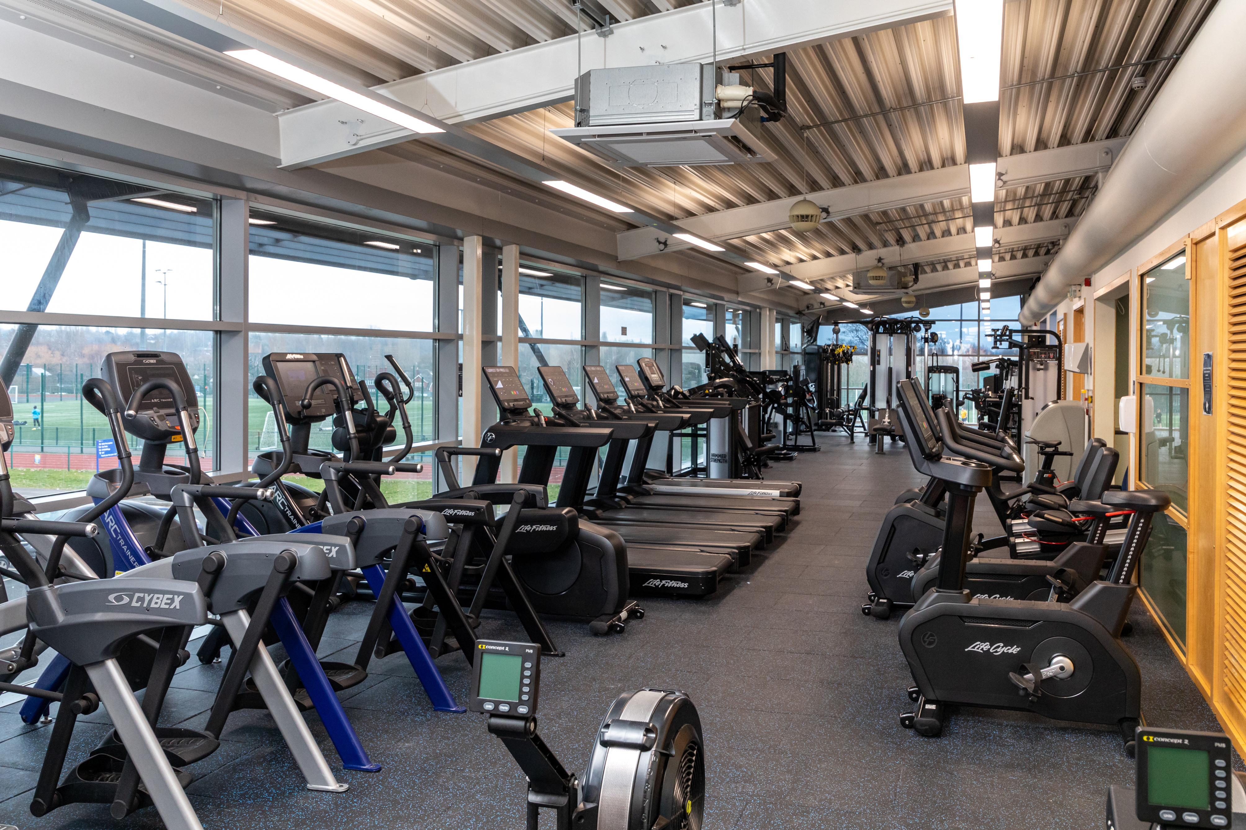 Exercise machines in the Walsall Campus gym, including rowing machines, weights, treadmills and bikes, a large window looking onto the outdoor sports facilities