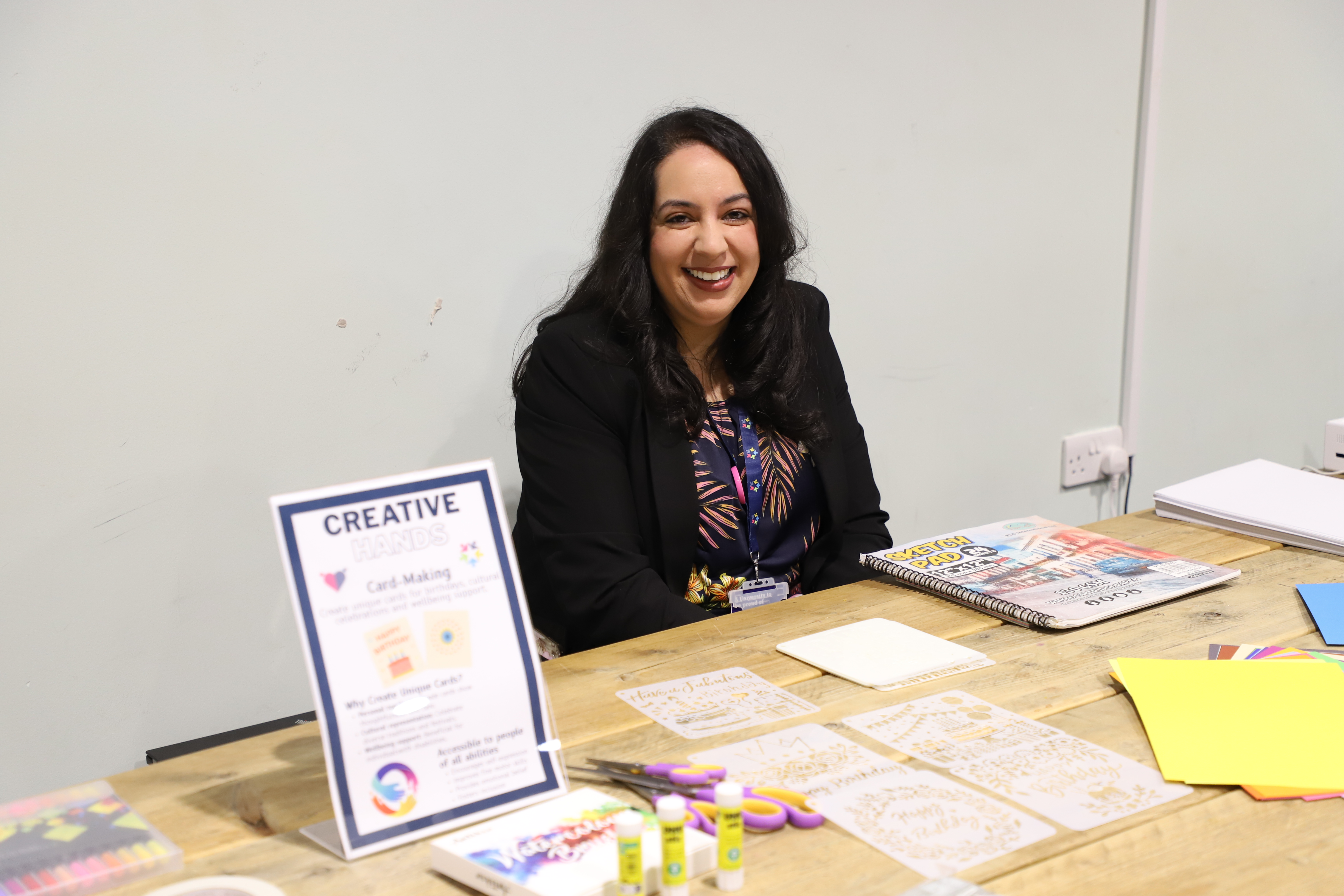 Lady with black hair, wearing a black blazer and colourful top, sitting at a desk. A sign stating creative hands. Arts and card making.