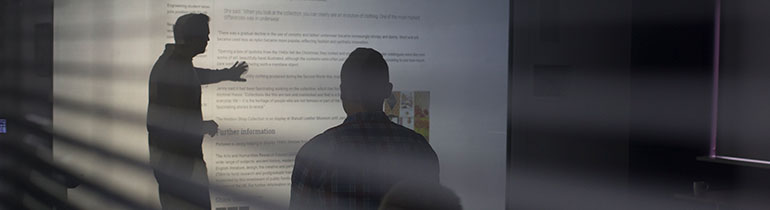 Shadows of a lecturer and student captured under the light of a projector in a dark room.
