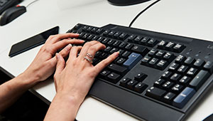 Photograph of a keyboard on a white table-top being operated by two hands of a person outside the frame.