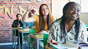 A student raising their hand in a classroom between other students sitting at their desks.