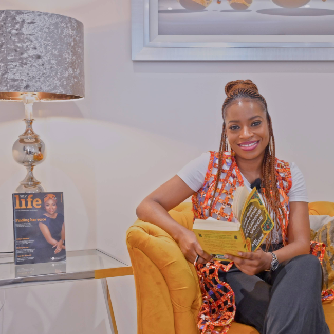 Abi Dare sitting in a yellow chair and reading next to a glass end table with a lamp